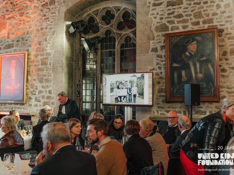 In einem historischen Saal mit rustikalen Steinwänden und großen Fenstern sind mehrere Personen an elegant gedeckten Tischen versammelt. An der Wand hängen zwei große Porträts, und zwei Scheinwerfer beleuchten die Szenerie. Im Vordergrund ist eine Gruppe von Menschen in Gespräche vertieft. Auf einem großen Bildschirm an der Wand ist ein Bild von mehreren Personen zu sehen. Unten rechts im Bild steht das Logo von "United Kids Foundations" mit dem Zusatz "20 Jahre".