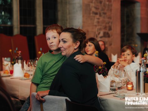 In einem festlich dekorierten Raum sitzen Personen an einem Tisch. Ein Kind in einem grünen T-Shirt lehnt sich an eine Frau, die liebevoll den Arm um das Kind gelegt hat. Im Hintergrund sitzen weitere Menschen an einem Tisch mit Blumenarrangement und Kerzen. Auf dem Tisch stehen Gläser und Wasserflaschen. Auf dem Bild ist das Logo der "United Kids Foundations" zu sehen.