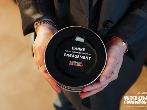 Eine Person in einem dunklen Mantel hält eine runde, offene Verpackung in den Händen. Darin befindet sich eine Auszeichnung mit der Aufschrift "DANKE FÜR IHR AUSSERGEWÖHNLICHES ENGAGEMENT" und dem Logo von "UNITED KIDS FOUNDATIONS" darauf. In der unteren rechten Ecke des Bildes steht "20 JAHRE UNITED KIDS FOUNDATIONS - Das Kindernetzwerk der BRAWO GROUP".