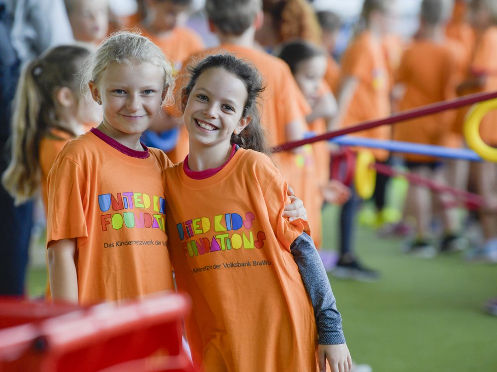 Zwei lachende Kinder in orangefarbenen T-Shirts bei einem Event. Im Hintergrund sind weitere Kinder.