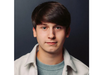 Ein junger Mann mit dunklem, kurzem Haar schaut direkt in die Kamera. Er trägt einen hellen Pullover und eine helle Jacke. Der Hintergrund ist dunkel und einfarbig, wodurch das Gesicht deutlich hervorsticht.