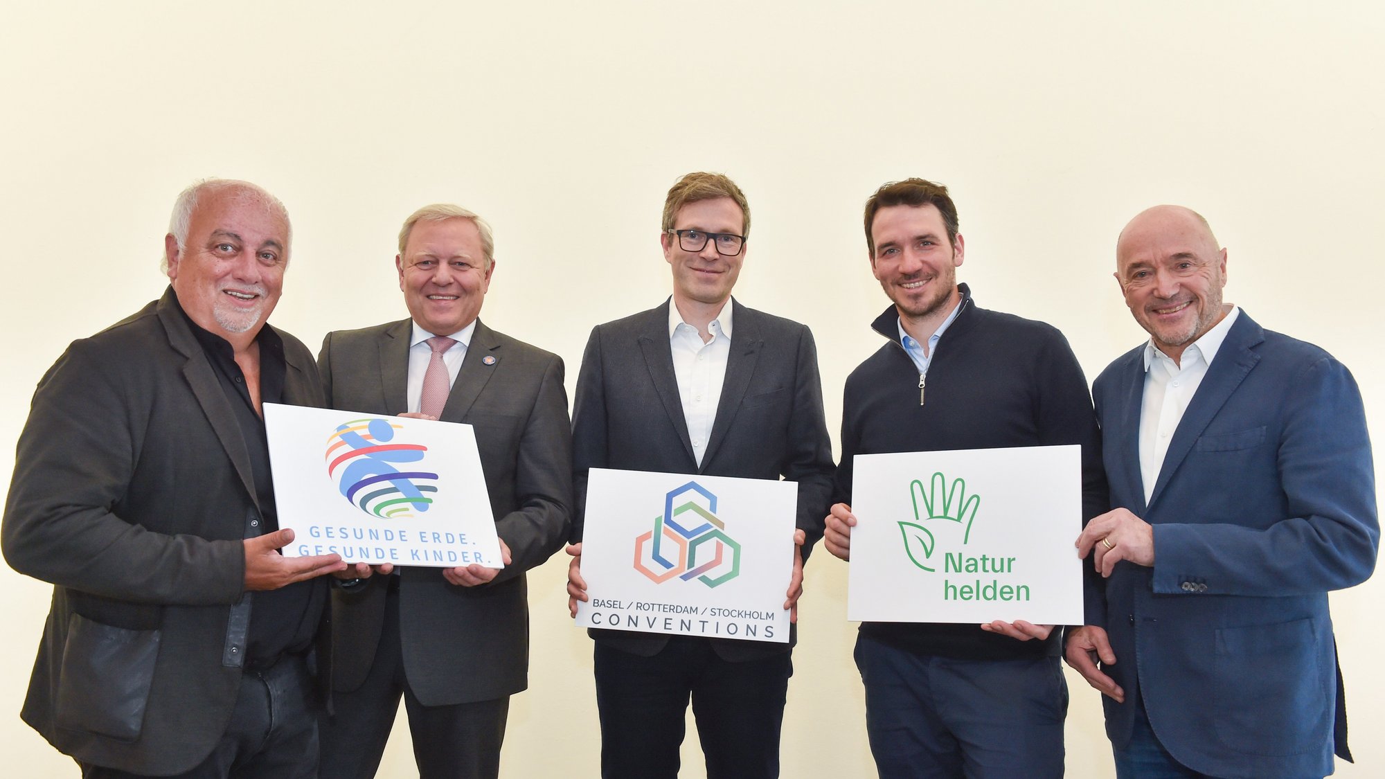 Fünf Personen im Anzug stehen lächelnd in einer Reihe. Sie halten drei Schilder mit verschiedenen Logos drauf vor ihre Brust.