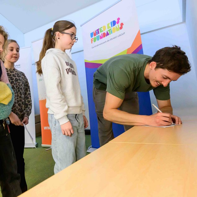 Eine Gruppe von Kindern steht in einer Reihe in einem Raum, während eine Person an einem Tisch etwas unterschreibt. Im Hintergrund sind bunte Plakate mit Text erkennbar. Die Kinder scheinen gespannt zu sein und warten geduldig. Der Raum wirkt hell und freundlich.