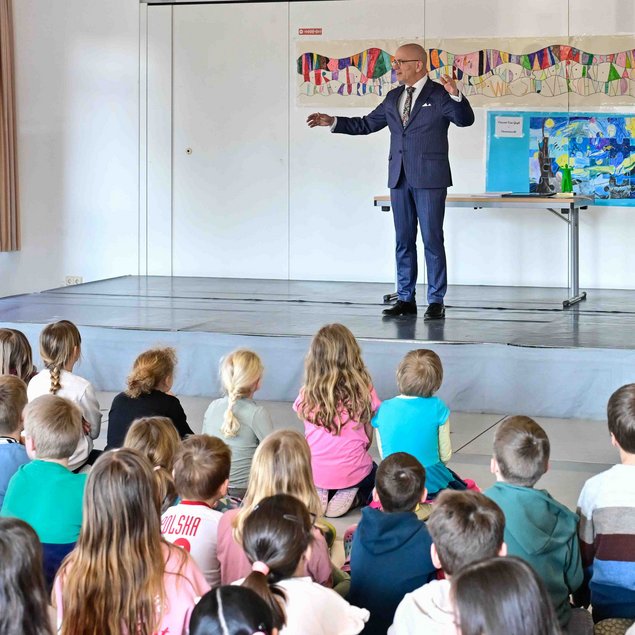 Ein Mann in einem Anzug steht auf einer kleinen Bühne und spricht zu einer Gruppe von Schulkindern, die auf dem Boden sitzen und aufmerksam zuhören. Im Hintergrund ist ein buntes Kunstwerk an der Wand zu sehen. Links steht eine Tafel mit mehreren Blättern darauf.