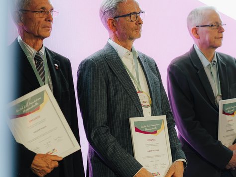 Drei ältere Männer stehen nebeneinander auf einer Bühne und halten Urkunden. Sie tragen Anzüge und wirken ernst. Im Hintergrund ist eine große, helle Leinwand. Auf der rechten unteren Seite des Bildes steht „20 Jahre United Kids Foundations - Das Kindernetzwerk der Brawo Group“.