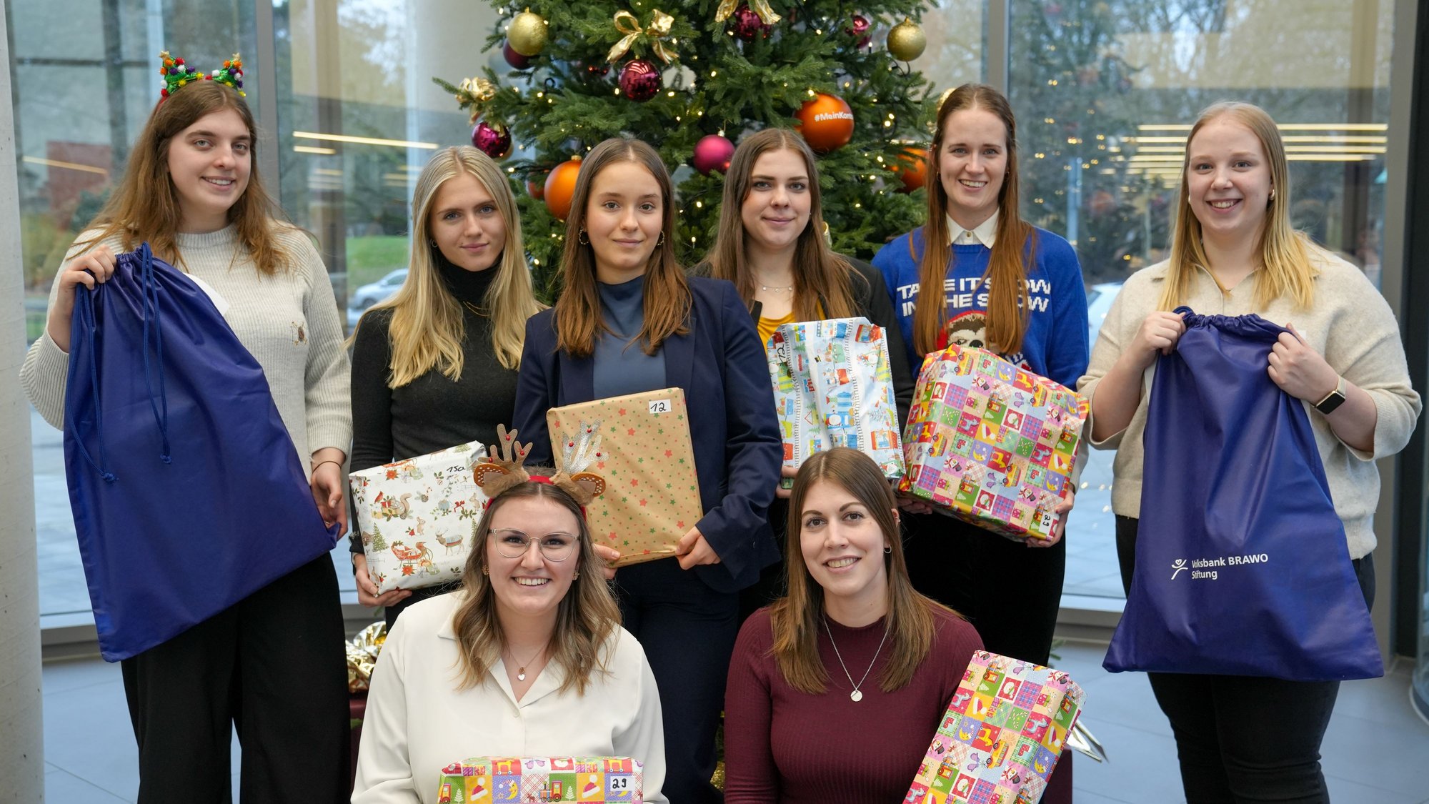 Fleißige Weihnachtsengel packten individuell zusammengestellte Geschenktüten, die bei den förderbedürftigen Kindern und Jugendlichen pünktlich unter dem Weihnachtsbaum liegen. Auf dem Bild sind acht Personen zu sehen. Sechs von ihnen stehend, zwei davor kniend. Hinter ihnen steht ein geschmückter Weihnachtsbaum. Die Personen haben zum Teil einen Sack und zum Teil bunt verpackte Geschenke in der Hand.