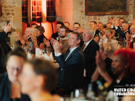 In einem festlich beleuchteten Raum sitzen viele elegant gekleidete Personen an runden Tischen, die mit Blumen und Gläsern dekoriert sind. Einige Personen klatschen, während ein Mann ein Foto mit seinem Smartphone macht. Die Atmosphäre wirkt freudig und konzentriert. Im Hintergrund sind Steinwände und bunte Lichteffekte zu sehen. Auf dem Bild steht "20 Jahre United Kids Foundations" in der Ecke.