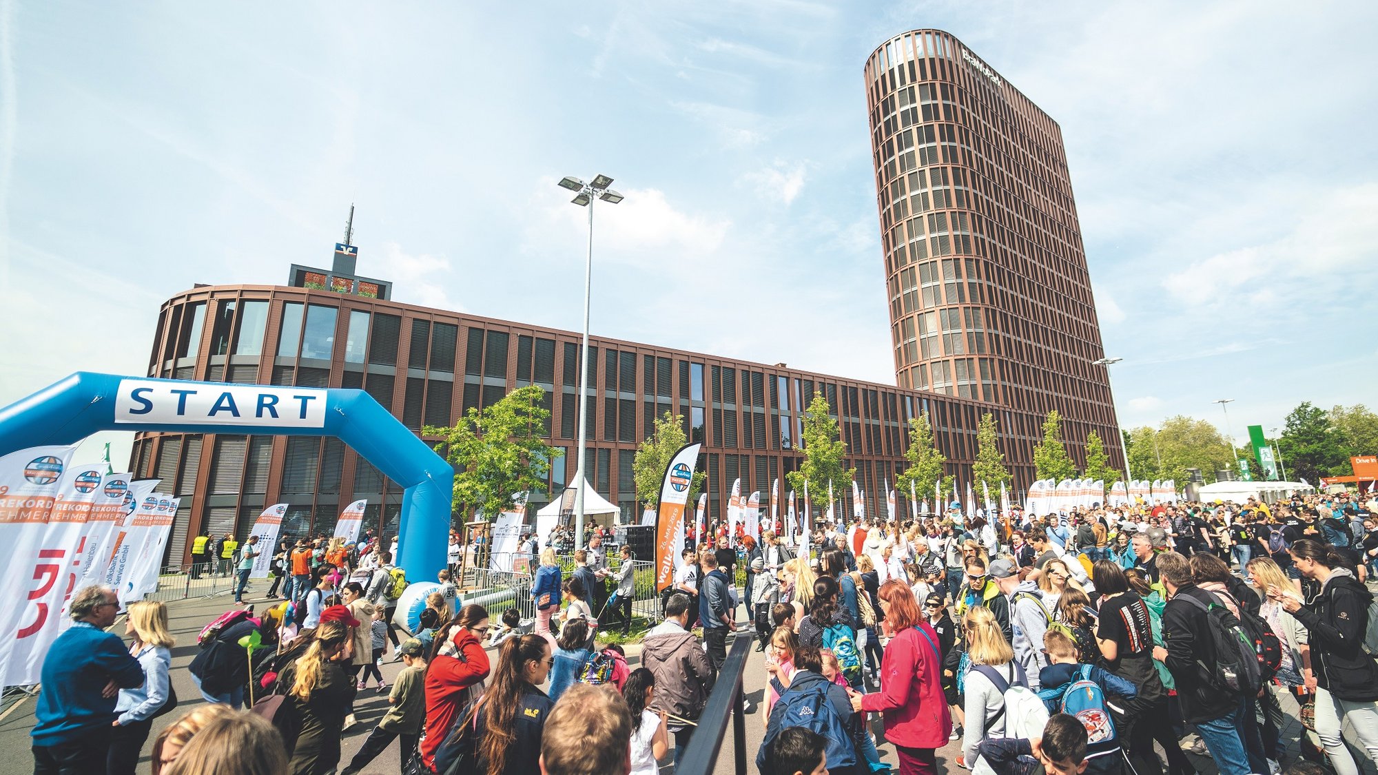 Tausende Menschen sammelten 2019 im Rahmen des walk4help Schritte und Spenden für Kinderhilfsprojekte. 2025 findet eine Neuauflage des Mega-Events im Braunschweiger Bürgerpark statt. Bildquelle: AUSDRUCKSLOS Auf dem Bild ist eine große Menschenmenge vor einem L-förmigen Gebäude zu sehen. Links im Bild ist ein aufblasbares, blaues Tor auf dem das Wort "Start" steht.