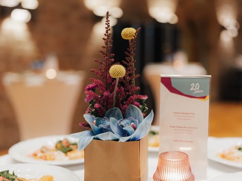 Ein festlich gedeckter Tisch in einem stimmungsvoll beleuchteten Raum. Im Vordergrund steht eine schlichte Papierbox als Vase mit einem bunten Blumenarrangement, bestehend aus gelben Kugelblumen, roten Zweigen und blauen Blüten. Daneben befindet sich ein Menükärtchen mit der Aufschrift "20 Jahre". Im Hintergrund sind mehrere Teller mit kunstvoll angerichteten Speisen zu sehen. Rechts steht ein Teelicht in einem geriffelten Glas. An einer Stelle im Bild steht "United Kids Foundations" und "Das Kindennetzwerk der Brawo Group“.