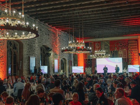 Das Bild zeigt einen festlich beleuchteten Saal mit zahlreichen Gästen, die an runden Tischen sitzen. Die Decke ist mit Holzbalken verziert und die Steinwände des Raums sind in warmem Rot angestrahlt. Im Hintergrund sind mehrere Bildschirme und ein Präsentationspodium zu sehen. Große Kronleuchter hängen von der Decke. Unten rechts im Bild ist das Logo der "United Kids Foundations" zu erkennen.