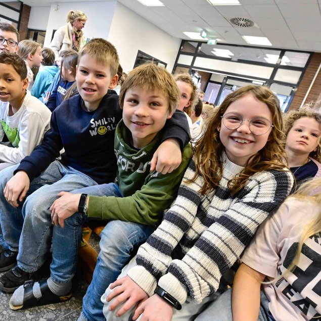 Auf dem Bild sind mehrere lachende Kinder zu sehen, die auf einer Bank in einem Innenraum sitzen. Einige tragen Brillen und sie sind lässig gekleidet. Im Hintergrund ist eine Frau mit einem Schal zu sehen, die steht und mit anderen Personen spricht. Die Umgebung wirkt wie ein Schulflur oder ein Gemeinschaftsraum. Die Kinder scheinen auf etwas zu warten oder an einer Veranstaltung teilzunehmen.