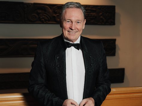 Ein älterer Mann steht lächelnd in einem formellen Ambiente. Er trägt einen eleganten schwarzen Anzug mit einer Fliege und ein weißes Hemd. Im Hintergrund sind kunstvoll verzierte Holzelemente und eine indirekte Beleuchtung zu sehen. Auf dem Bild ist unten rechts das Logo mit der Aufschrift "20 Jahre United Kids Foundations" zu erkennen.