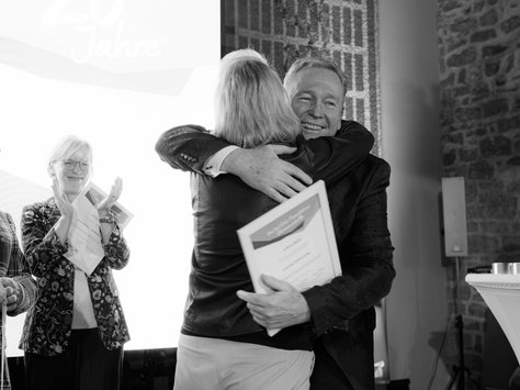 Zwei Personen umarmen sich herzlich bei einer Feier. Die Person links trägt ein Zertifikat in den Händen, während eine weitere Person in festlicher Kleidung applaudiert. Im Hintergrund ist ein leuchtendes Schild mit der Aufschrift "20 Jahre" sichtbar. Rechts unten im Bild steht „United Kids Foundations“.