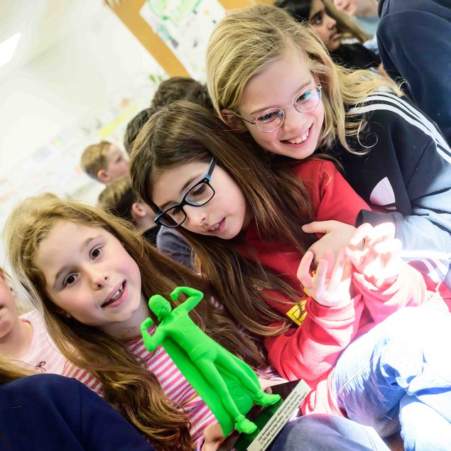 Eine Gruppe junger Mädchen sitzt dicht beieinander auf einem Stuhl. Zwei Mädchen in der Mitte schauen auf etwas, das ein Mädchen hält: eine grüne, muskulöse Figur. Die Atmosphäre wirkt fröhlich und lebhaft, umgeben von weiteren Kindern im Hintergrund.