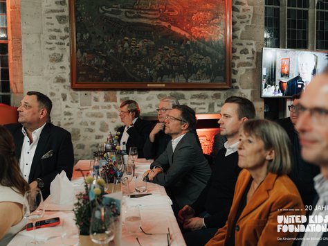 Eine Gruppe von Personen sitzt an festlich gedeckten Tischen in einem Raum mit Steinwänden und großen Fenstern. Im Hintergrund hängt ein großes Gemälde, und ein Bildschirm zeigt das Bild einer Person. Die Tische sind mit Blumen und Gläsern dekoriert. In der unteren rechten Ecke steht "20 Jahre United Kids Foundations – Das Kindernetzwerk der Brawo Group".