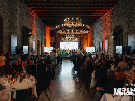 In einem großen, stimmungsvoll beleuchteten Saal sitzen zahlreiche Menschen an runden Tischen, die festlich gedeckt sind. Im Vordergrund sind gedeckte Tische mit weißen Tischdecken, Kerzen und Gläsern zu sehen. An der Stirnseite des Raumes gibt es eine beleuchtete Bühne, auf der eine Gruppe von Kindern in farbenfroher Kleidung steht. Große Kronleuchter hängen von der Decke und tauchen den Raum in warmes Licht. Die Wände sind aus Stein mit gerahmten Kunstwerken. Monitore zeigen das Logo des "United Kids Foundations"-Events zum 20-jährigen Jubiläum.