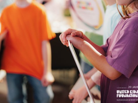 Das Bild zeigt mehrere Kinder in bunten T-Shirts, die in einer Gruppe stehen. Eines der Kinder hält ein langes, stabförmiges Objekt. Im Hintergrund sind unscharfe Formen und Farben erkennbar. In der unteren rechten Ecke ist das Logo von "United Kids Foundations" und der Hinweis "20 Jahre" zu sehen.