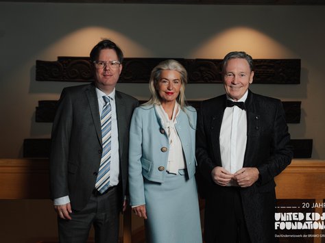 Drei Personen stehen nebeneinander und lächeln in die Kamera. Links ist ein Mann in einem grauen Anzug mit gestreifter Krawatte. In der Mitte steht eine Frau in einem hellblauen Kostüm mit passender Jacke und weißer Bluse. Rechts ist ein Mann in einem schwarzen Anzug mit weißem Hemd und Fliege. Im Hintergrund sind dekorative Holzverzierungen an der Wand zu sehen. Unten rechts im Bild befindet sich das Logo von "United Kids Foundations" mit dem Hinweis "20 Jahre".