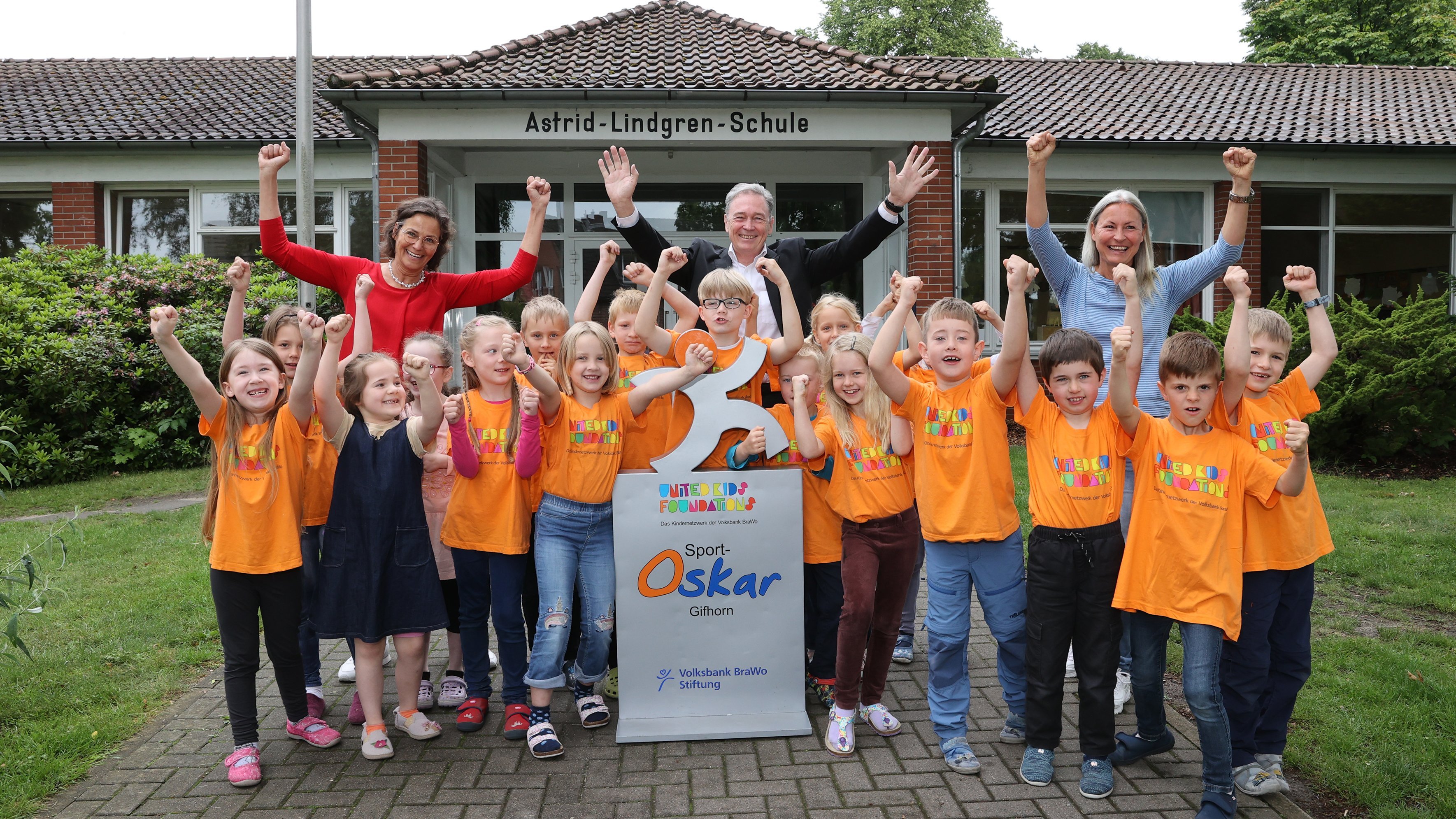 Eine Gruppe aus Kindern in orangenen Tshirts, sowie vereinzelte Erwachsene stehen vor einem Gebäude mit der Aufschrift "Astrid-Lindgren-Schule" und strecken ihre Arme freudig nach oben.