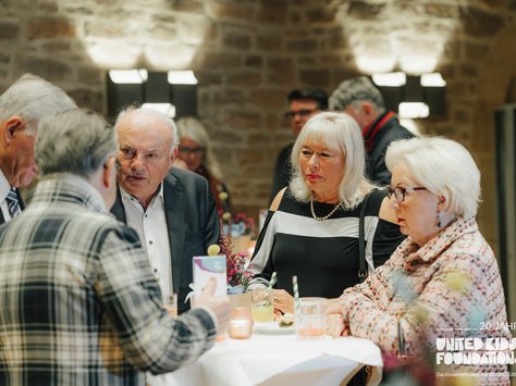 In einem gemütlich beleuchteten Raum stehen mehrere Personen um einen runden Stehtisch und unterhalten sich. Einige trinken aus Gläsern, die auf dem Tisch stehen. Die Gruppe, bestehend aus Männern und Frauen, scheint in ein angeregtes Gespräch vertieft zu sein. Im Hintergrund sind weitere Personen und eine gemauerte Wand sichtbar. Auf dem Bild ist ein Logo mit der Aufschrift "20 Jahre United Kids Foundations" zu erkennen.