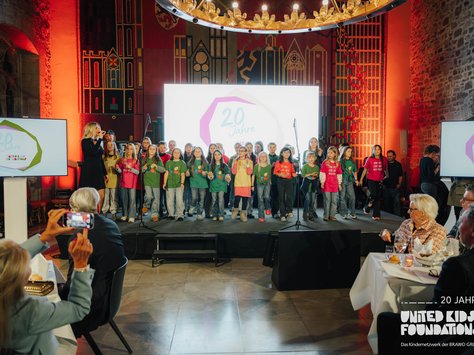 Eine Gruppe von Kindern steht auf einer festlich beleuchteten Bühne und singt. Die Kinder tragen bunte T-Shirts. Links steht eine Frau mit einem Mikrofon, die mit ihnen singt. Im Hintergrund ist ein großes Display mit der Aufschrift "20 Jahre" zu sehen. Im Vordergrund sitzen Zuschauer an Tischen und eine Person hält ein Smartphone in die Luft, um ein Foto oder Video zu machen. Die Umgebung wirkt festlich und der Raum ist mit Lampen und Dekorationen geschmückt.