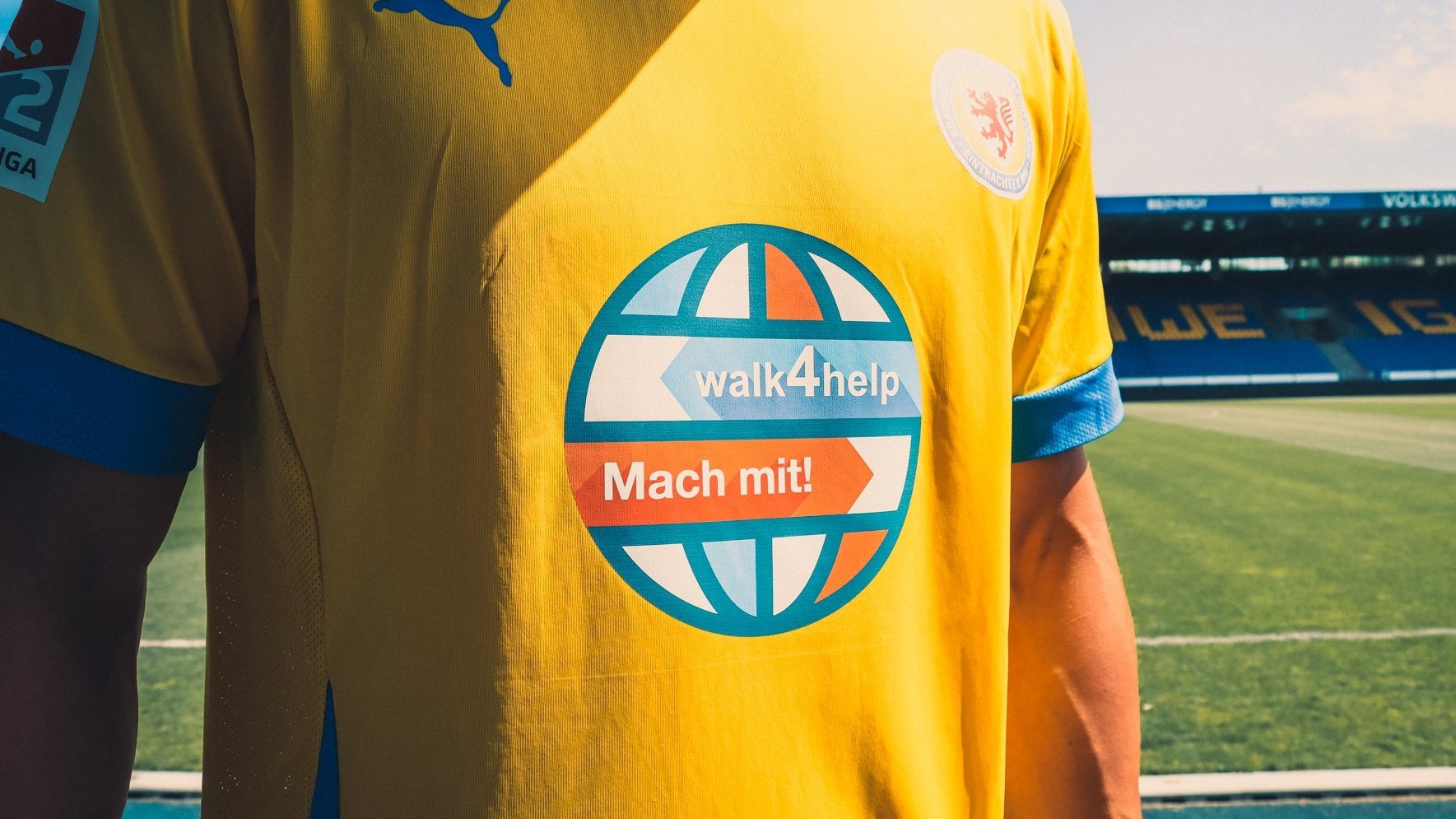 Nahaufnahme des gelben Fußballtrikots eines Spielers im Stadion. Im Zentrum ist das runde Sponsorenlogo "walk4help – Mach mit!" gut sichtbar, darüber das Puma-Logo und das Vereinswappen von Eintracht Braunschweig. Im Hintergrund ist das leere Fußballfeld und ein Teil der Tribüne zu sehen.