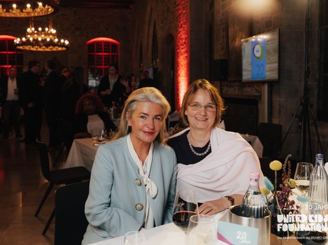 Zwei Frauen sitzen an einem festlich gedeckten Tisch in einem elegant beleuchteten Raum. Die Frau links trägt einen hellblauen Blazer, die Frau rechts ist in ein rosa Tuch gewickelt. Auf dem Tisch stehen Gläser mit Getränken und Blumendekorationen. Im Hintergrund sind weitere Gäste und ein Bildschirm zu erkennen. An der Ecke des Bildes ist das Logo der "United Kids Foundations" mit dem Hinweis auf das 20-jährige Bestehen zu sehen.