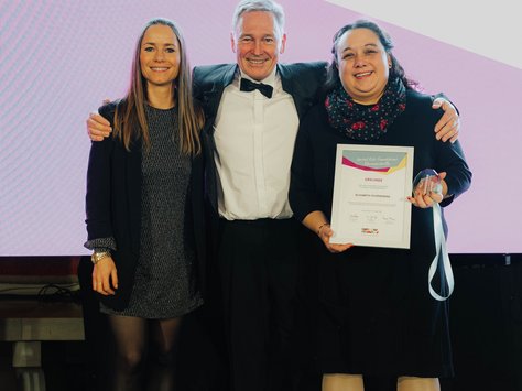 Das Bild zeigt drei Personen, die zusammen auf einer Bühne stehen. Die Person in der Mitte trägt einen dunklen Anzug mit Fliege und hat ihre Arme um die beiden anderen Personen gelegt. Die Person rechts hält eine Urkunde und eine Medaille in der Hand. Links ist das Logo der "United Kids Foundations" zu sehen. Der Hintergrund ist hell beleuchtet, und es scheint sich um eine Preisverleihung oder ein besonderes Event zu handeln.