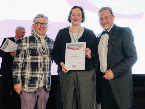 Drei Personen stehen vor einem großen Bildschirm, auf dem "20 Jahre United Kids Foundations" angezeigt wird. Die Person in der Mitte hält eine Urkunde in der Hand. Links steht eine Person mit einem karierten Sakko und rechts eine Person im schwarzen Anzug. Alle drei lächeln in die Kamera. Im Hintergrund sind weitere Personen zu sehen.