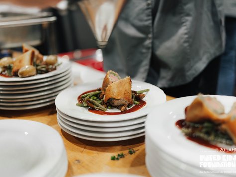 Ein Tisch, auf dem mehrere gestapelte weiße Teller stehen. Auf den vordersten Tellern sind kunstvoll angerichtete Speisen zu sehen: Gebratene Stücke, möglicherweise Fleisch, umgeben von einer Sauce und grünen Bohnen. Im Hintergrund ist ein Mensch in grauer Kleidung zu erkennen, der mit einer Küchentechnik beschäftigt ist. Auf dem Bildrand ist ein Schriftzug zu „20 Jahre United Kids Foundations“ sichtbar.