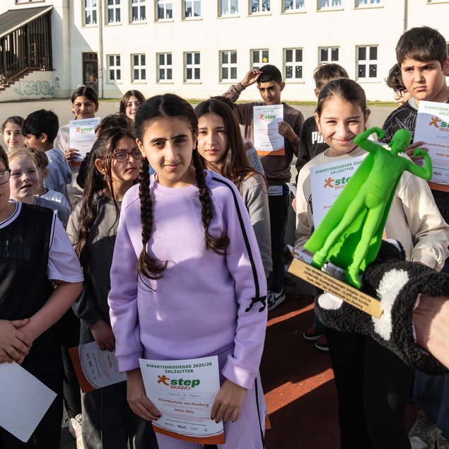 Eine Gruppe von Schülern steht draußen vor einem Schulgebäude und hält Urkunden in den Händen. Im Vordergrund ist ein Mädchen mit Zöpfen, das eine Urkunde mit dem Logo "step BRAVO" trägt. Jemand im Vordergrund hält eine grüne Plastikfigur, die wie ein Pokal aussieht. Die Schüler wirken stolz und glücklich. Im Hintergrund ist ein Schulgebäude mit mehreren Fenstern zu sehen.