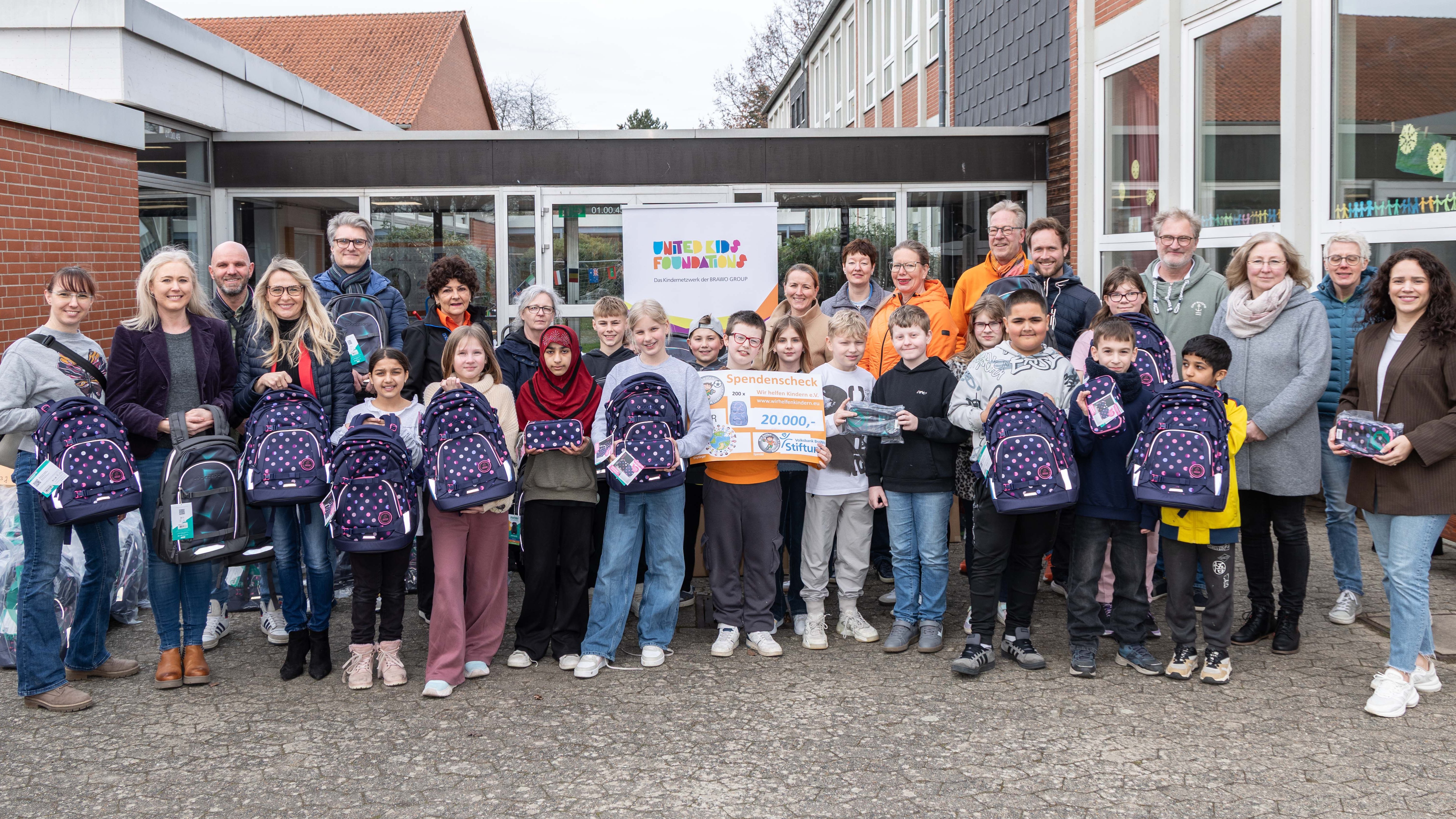 Eine große Gruppe von Kindern und Erwachsenen steht vor einem Schulgebäude. Die Kinder halten neue Schultaschen mit einem Punktmuster in den Händen. Eine Person hält einen großen Spendenscheck über 20.000 Euro. Im Hintergrund ist ein Banner mit der Aufschrift "United Kids Foundations" sichtbar. Alle wirken zufrieden und fröhlich.
