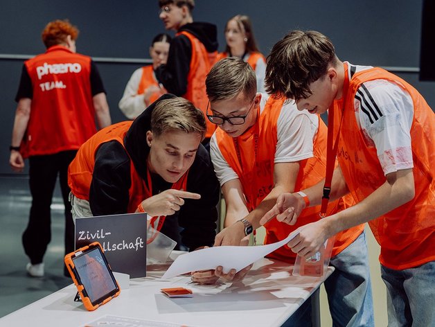 Drei Jugendliche die über einen Tisch gelehnt stehen und nachdenklich auf ein Blatt Papier schauen. Im Hintergrund sind leicht unscharf, ebenfalls Jugendliche zu sehen. Alle tragen ein orangenes Leibchen. Auf dem Tisch im Vordergrund steht zudem seitlich ein Tablet mit orangener Hülle, sowie ein Schild mit der Aufschrift "Zivilgesellschaft".
