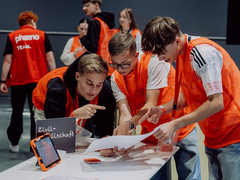 Drei Jugendliche die über einen Tisch gelehnt stehen und nachdenklich auf ein Blatt Papier schauen. Im Hintergrund sind leicht unscharf, ebenfalls Jugendliche zu sehen. Alle tragen ein orangenes Leibchen. Auf dem Tisch im Vordergrund steht zudem seitlich ein Tablet mit orangener Hülle, sowie ein Schild mit der Aufschrift "Zivilgesellschaft".