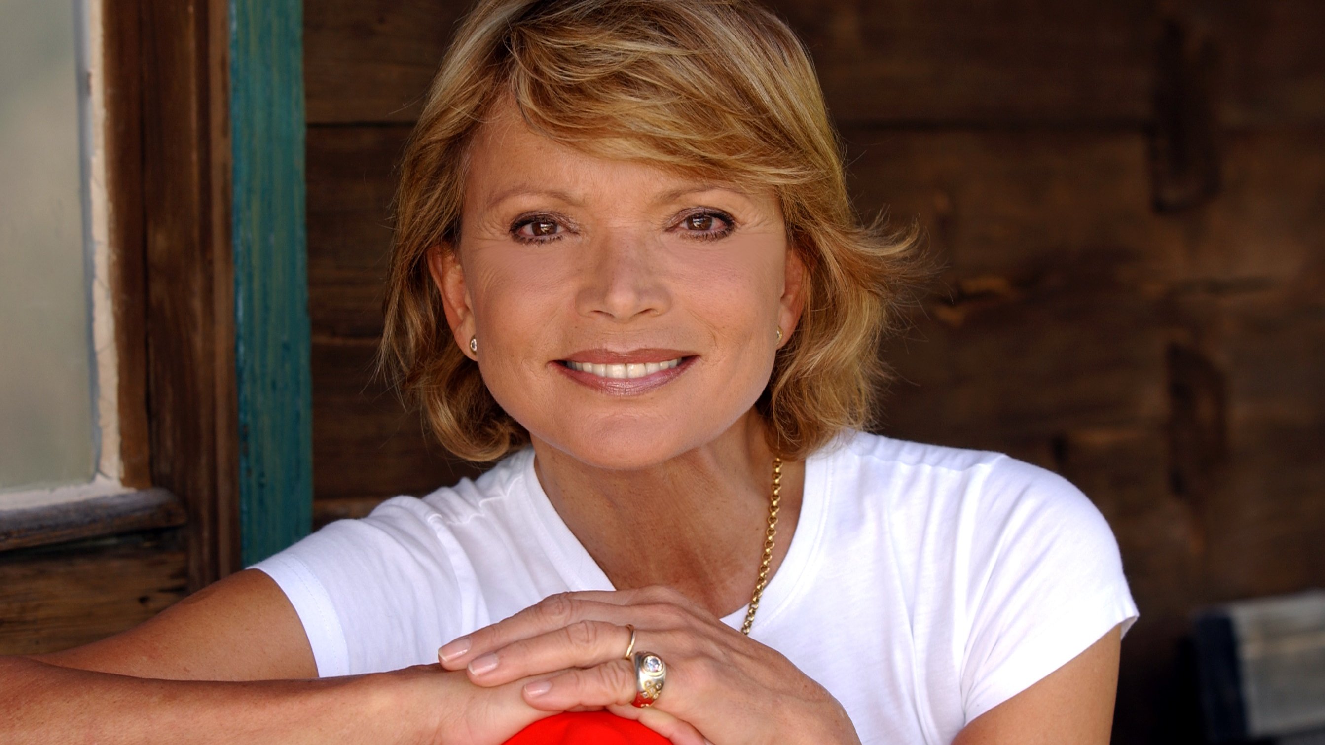 Eine lächelnde Person mit kurzen, blonden Haaren sitzt vor einer Holzwand. Sie trägt ein weißes T-Shirt und hat die Arme verschränkt. Ein goldener Ring und eine Kette sind sichtbar.