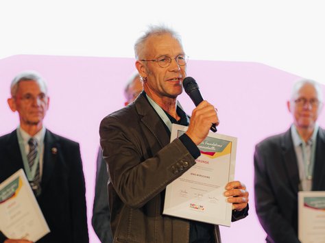 Ein älterer Mann steht auf einer Bühne vor einem Mikrofon und hält ein gerahmtes Zertifikat in der Hand. Hinter ihm stehen mehrere Männer in Business-Kleidung, die ebenfalls Zertifikate tragen. Im Hintergrund ist ein großer Bildschirm mit dem Logo der "United Kids Foundations" zu sehen. Der Anlass scheint eine Feier oder Ehrung zu sein, wie das auf dem Bildschirm angekündigte 20-jährige Jubiläum der Stiftung nahelegt.