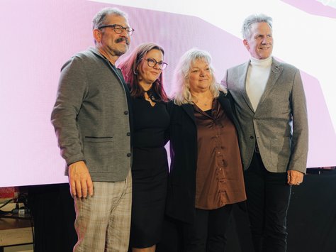 Vier Personen stehen auf einer Bühne nebeneinander und posieren für ein Foto. Sie sind schick gekleidet, drei tragen Jacken, während eine Person ein schwarzes Kleid trägt. Der Hintergrund ist eine große, bunte Leinwand, auf der das Logo von "United Kids Foundations" und der Schriftzug "20 Jahre" zu sehen sind. Es handelt sich um eine Veranstaltung, die das 20-jährige Jubiläum der Stiftung feiert.