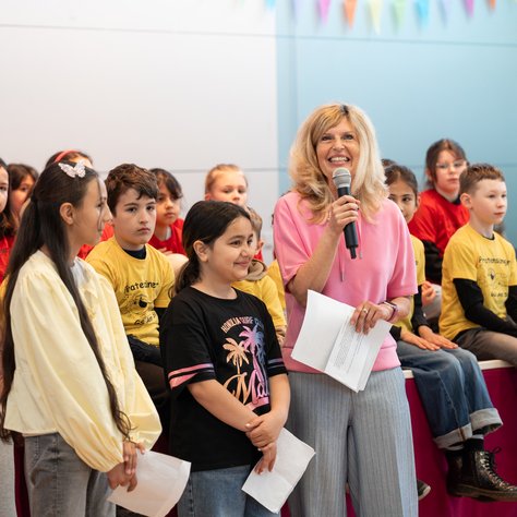 Eine Frau in einem rosa Pullover hält ein Mikrofon und spricht zu einer Gruppe von Kindern, die auf Bänken sitzen. Die Kinder tragen bunte T-Shirts, einige in Rot und andere in Gelb. Zwei Mädchen stehen neben der Frau und halten Papiere in der Hand. Im Hintergrund sind bunte Wimpel zu sehen, die eine fröhliche Atmosphäre schaffen.