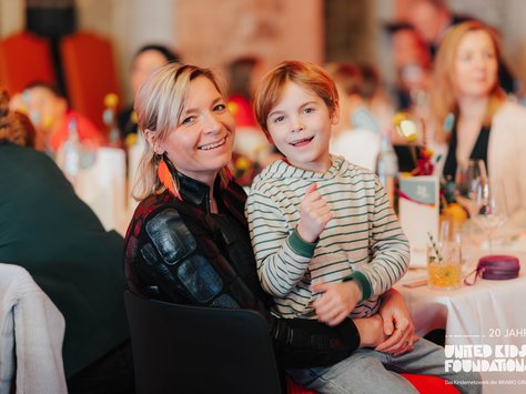 Eine Frau und ein Kind sitzen lächelnd an einem Tisch in einem festlich dekorierten Raum. Die Frau trägt eine Jacke mit auffälligen Mustern und orangene Ohrringe, während das Kind einen gestreiften Pullover trägt. Im Hintergrund sind weitere Personen an Tischen zu sehen. Auf dem Bild ist das Logo der "United Kids Foundations - 20 Jahre" zu erkennen.
