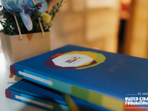 Zwei blaue Bücher mit einem bunten Aufdruck liegen gestapelt auf einem Tisch. Im Hintergrund befindet sich eine kleine Tasche mit Blumen in verschiedenen Farben. Links unten im Bild ist das "United Kids Foundations" Logo und der Schriftzug "20 Jahre" zu sehen.