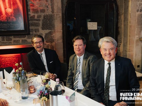 Drei Männer sitzen an einem elegant gedeckten Tisch in einem Raum mit Steinwänden. Der Tisch ist mit Gläsern, Blumen und Wasserflaschen dekoriert. Links ist ein Gemälde an der Wand zu sehen. Alle drei Männer tragen Anzüge und lächeln in die Kamera. Auf der rechten unteren Seite befindet sich das Logo der "United Kids Foundations" mit einem Hinweis auf das 20-jährige Jubiläum.