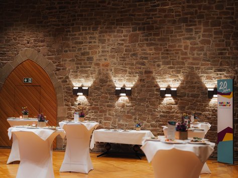 In einem eleganten Raum mit einer Steinwand stehen mehrere Stehtische, die mit weißen Tischdecken bedeckt sind. Jeder Tisch ist mit kleinen Blumengestecken geschmückt. Im Hintergrund sieht man eine große hölzerne Tür mit einem Notausgangsschild darüber. An der Wand hängen moderne Wandleuchten. Auf der rechten Seite befindet sich ein Roll-Up-Banner mit der Aufschrift "20 Jahre United Kids Foundations". Der Holzboden verleiht dem Raum eine warme Atmosphäre.