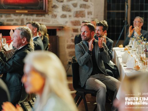 Das Bild zeigt eine Gruppe von Menschen, die an einem elegant gedeckten Tisch sitzen. Sie applaudieren, während sie aufmerksam nach vorne blicken. Im Vordergrund befindet sich eine Person, die unscharf ist. Der Raum hat eine warme, einladende Atmosphäre mit einer Steinwand im Hintergrund. In der rechten unteren Ecke des Bildes ist das Logo von "United Kids Foundations" mit der Aufschrift "20 Jahre" zu sehen.