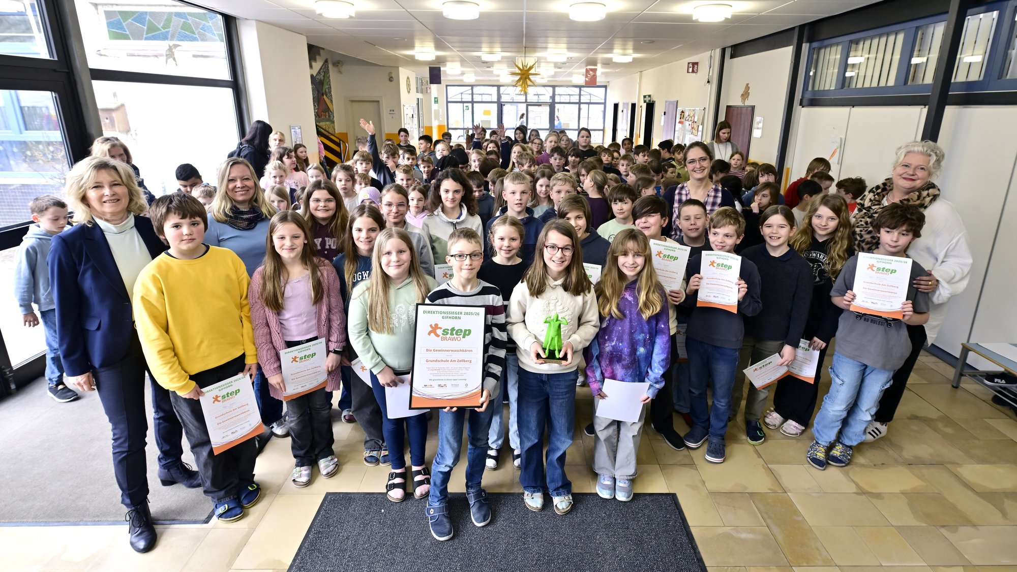In einem hell erleuchteten Schulflur steht eine große Gruppe von Kindern zusammen mit einigen Erwachsenen. Die Kinder sind überwiegend im mittleren Schulalter und halten stolz Urkunden in den Händen. Ein Mädchen in der vorderen Reihe hält eine grüne Trophy. Im Hintergrund ist eine festliche Dekoration zu sehen, die auf ein besonderes Ereignis oder eine Preisverleihung hindeutet. Die Atmosphäre wirkt fröhlich und feierlich.