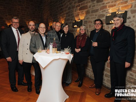 Eine Gruppe von acht Personen steht in Abendgarderobe um einen Hochtisch in einem festlich beleuchteten Raum mit Backsteinmauern. Auf dem Tisch befinden sich Getränke und Snacks. Die Personen lächeln in die Kamera. Im Hintergrund erkennt man weitere Gäste. Unten rechts im Bild ist ein Logo mit der Aufschrift "20 Jahre United Kids Foundations".