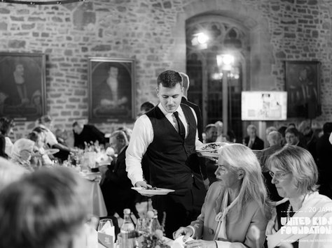 Das Bild zeigt eine elegante Veranstaltung in einem historischen Raum mit freiliegenden Steinmauern und großen Fenstern. Ein Kellner im Anzug serviert einem Gast am Tisch ein Gericht. Die Tische sind festlich gedeckt, umgeben von zahlreichen Gästen, die in Gesprächen vertieft sind. Im Hintergrund hängen mehrere Porträtgemälde an der Wand. Das gesamte Ambiente wirkt stilvoll und gehoben. Unten rechts ist das Logo der "United Kids Foundations" zu sehen.