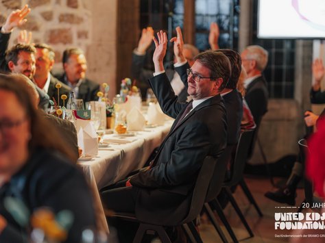 In einem festlichen Raum sitzen mehrere Leute an einem Tisch, der schön gedeckt ist. Einige Personen haben ihre Hände in die Luft gehoben, als würden sie abstimmen oder eine Frage beantworten. Auf dem Tisch stehen gefaltete Servietten, Wasserflaschen und Blumendekorationen. Am rechten unteren Bildrand ist das Logo von "United Kids Foundations" zu sehen, das ein 20-jähriges Jubiläum feiert. Die Stimmung wirkt engagiert und feierlich.