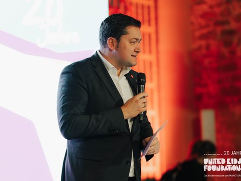 Ein Mann in einem dunklen Anzug spricht mit einem Mikrofon in der Hand. Er steht vor einem Hintergrund, der in warmen Rottönen beleuchtet ist. Im Hintergrund ist teilweise ein Steinmuster zu erkennen. Auf der linken Seite des Bildes ist ein unscharfer Schriftzug zu sehen, der "20 Jahre" andeutet. Unten rechts im Bild befindet sich das Logo der "United Kids Foundations" mit dem Zusatz "Das Kindernetzwerk der BRAVO GROUP".