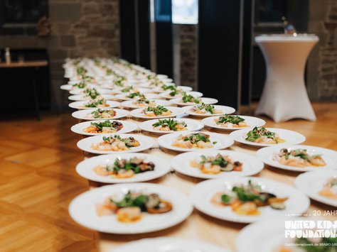 Ein langer Tisch ist mit vielen Tellern gedeckt, die sorgfältig angerichtetes Essen mit Gemüse und Fleisch enthalten. Die Teller sind in mehreren Reihen angeordnet und ziehen sich bis zum hinteren Bereich des Raumes. Der Boden ist aus hellem Holz und im Hintergrund sind Steinwände und ein rundes Stehtischchen sichtbar. In der Ecke ist das Logo "United Kids Foundations" und ein Hinweis auf das 20-jährige Jubiläum.