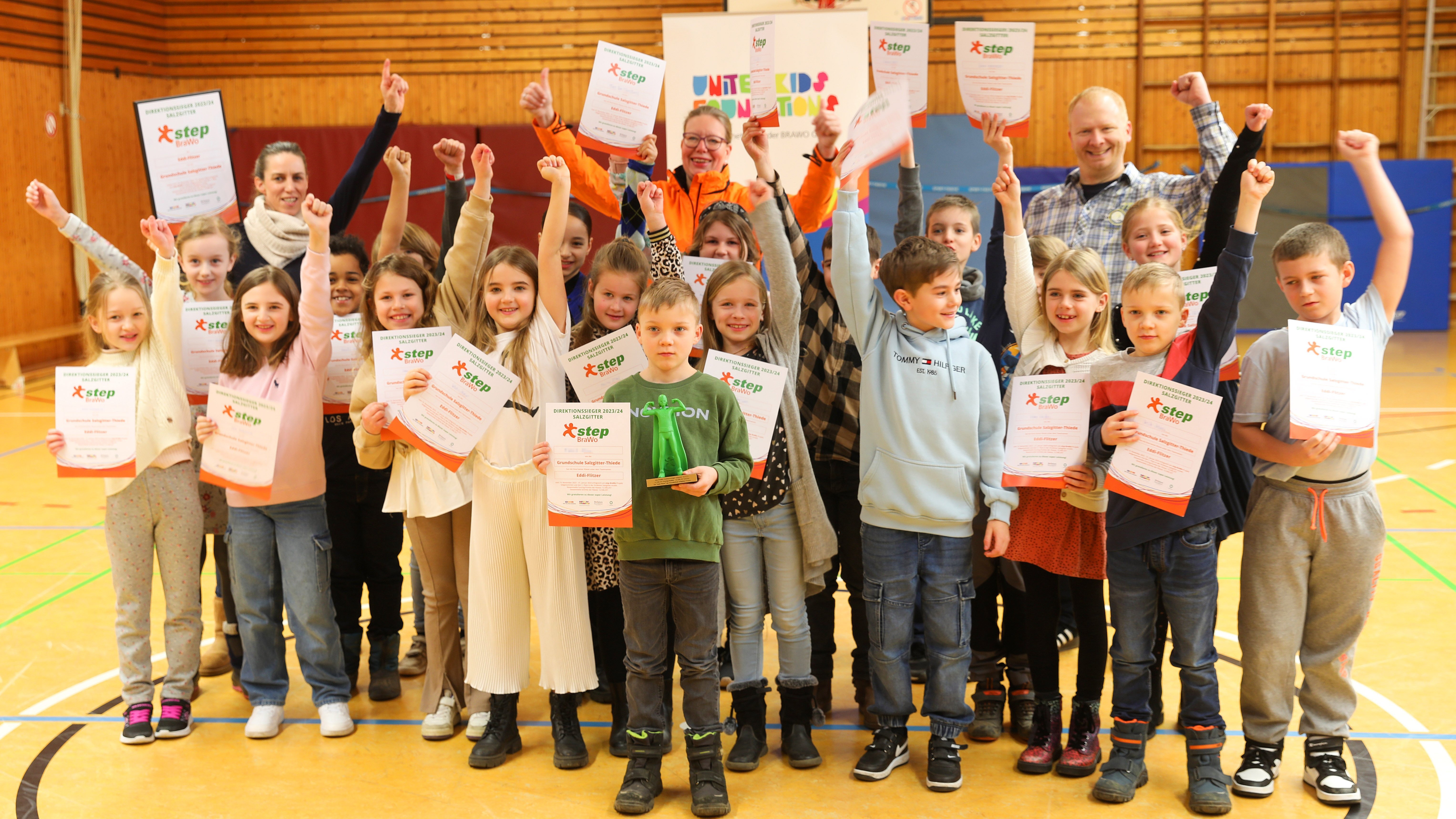 Die Kinder freuen sich über ihre Siegerurkunden, 250 Euro Preisgeld für die Klassenkasse und den Pokal. Auf dem Bild ist eine Gruppe aus Kindern und zwei Erwachsene zu sehen, die in einer Sporthalle stehen und ihre Arme freudig nach oben strecken. Sie halten alle eine Urkunde in der Hand.